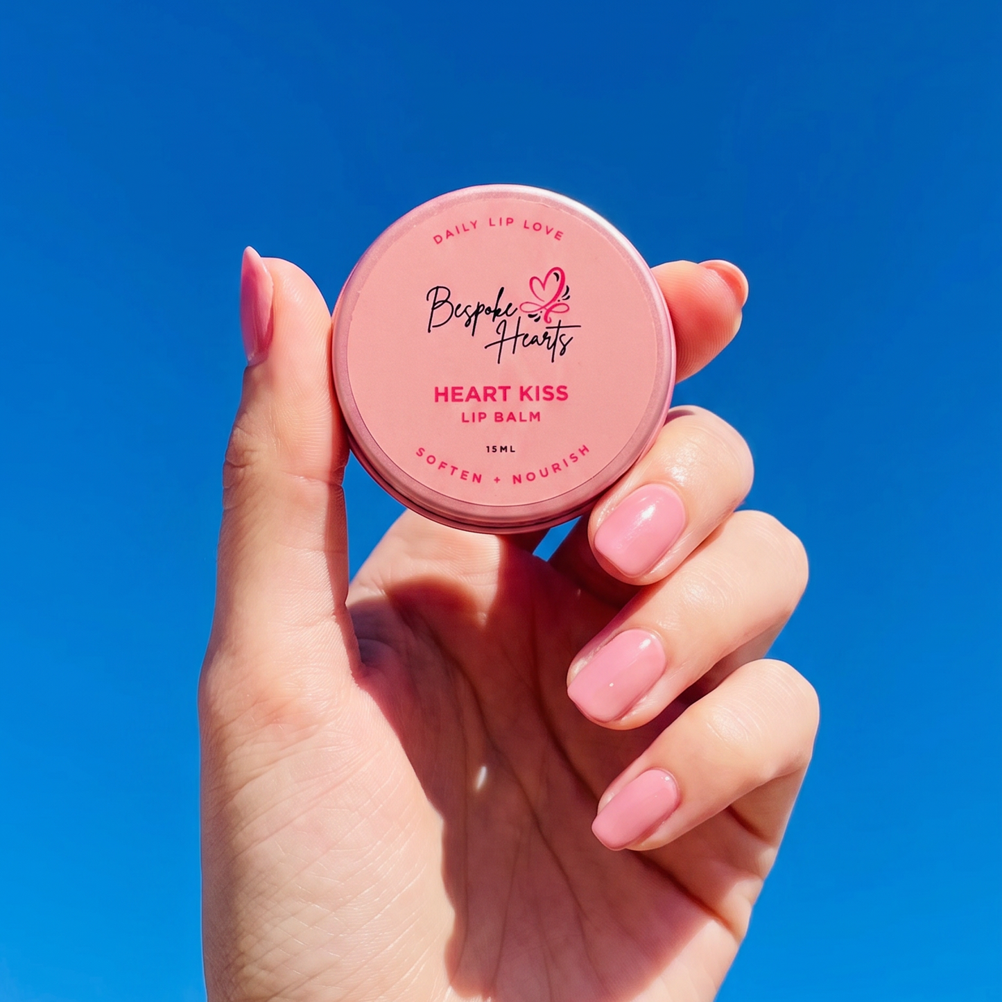 A hand with pink manicured nails holds a round pink compact of Heart Kiss Lip Balm against a bright blue sky back