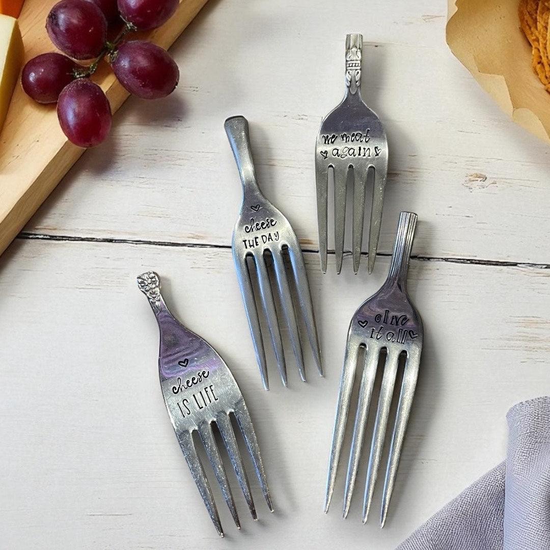 Set of four decorative forks with engraved text on a wooden surface with grapes and cheese.