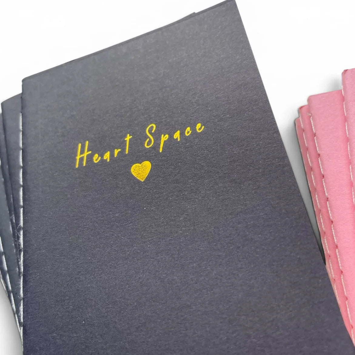 Black notebook with gold 'Heart Space' text and heart, alongside pink stitched notebook