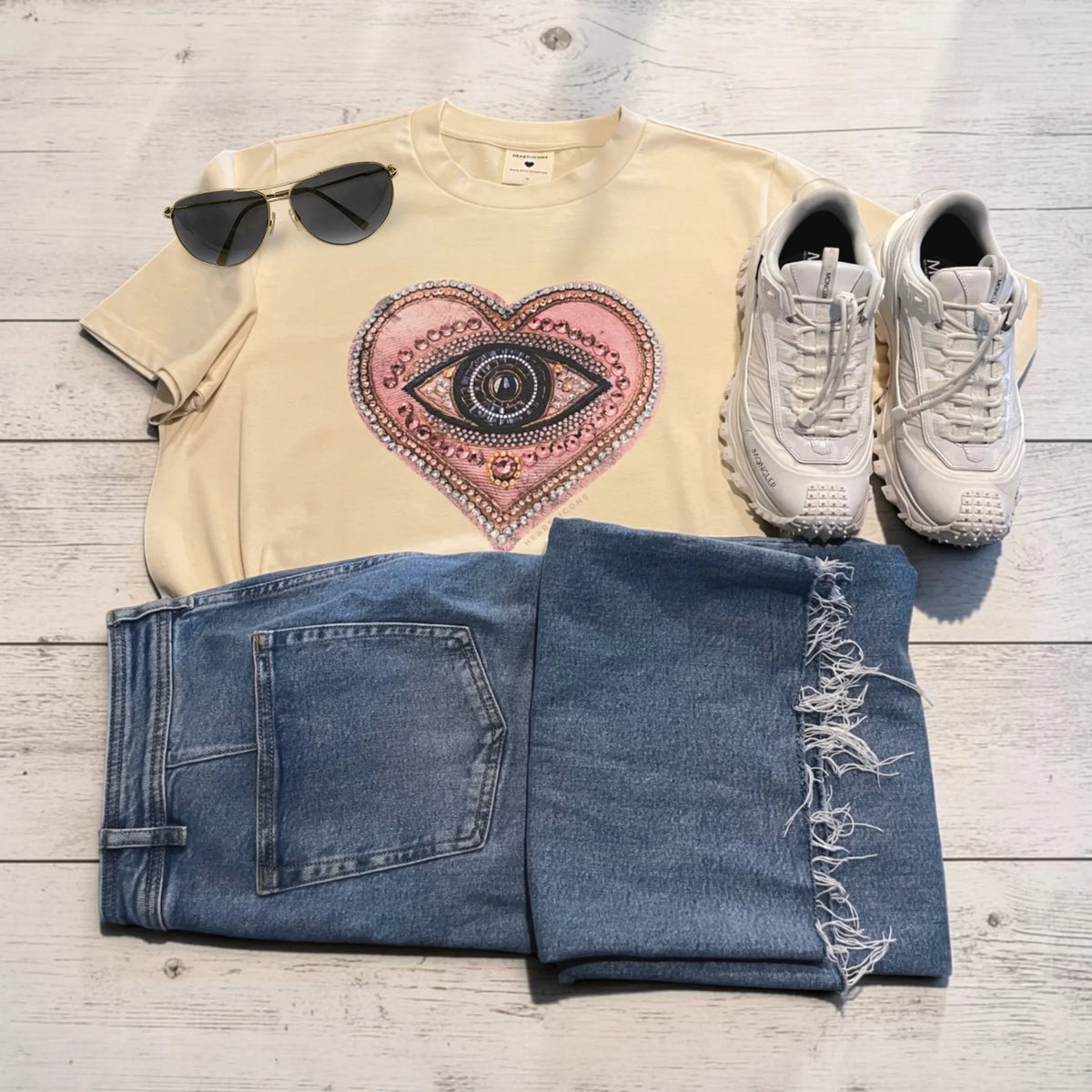 A styled flat-lay outfit on a light wood background featuring a cream t-shirt with a pink and black eye-in-heart graphic, blue jeans with frayed hems, white sneakers, and black sunglasses.
