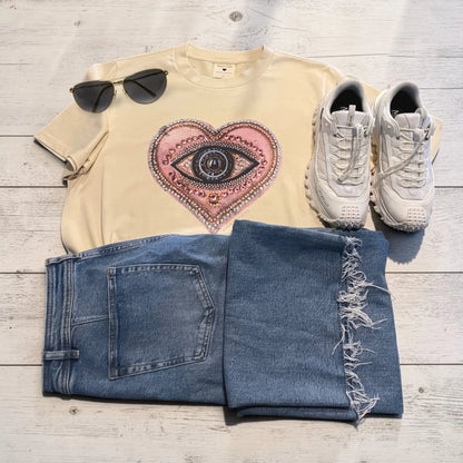 A styled flat-lay outfit on a light wood background featuring a cream t-shirt with a pink and black eye-in-heart graphic, blue jeans with frayed hems, white sneakers, and black sunglasses.