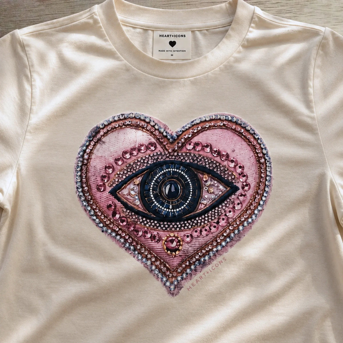 A close-up of a cream-colored t-shirt featuring a large, detailed graphic of a heart-shaped eye with pink, purple, and blue tones, embellished with rhinestone-like patterns. The brand name 'HEARTICONS' is visible on the inner neck label.