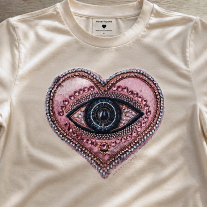 A close-up of a cream-colored t-shirt featuring a large, detailed graphic of a heart-shaped eye with pink, purple, and blue tones, embellished with rhinestone-like patterns. The brand name 'HEARTICONS' is visible on the inner neck label.