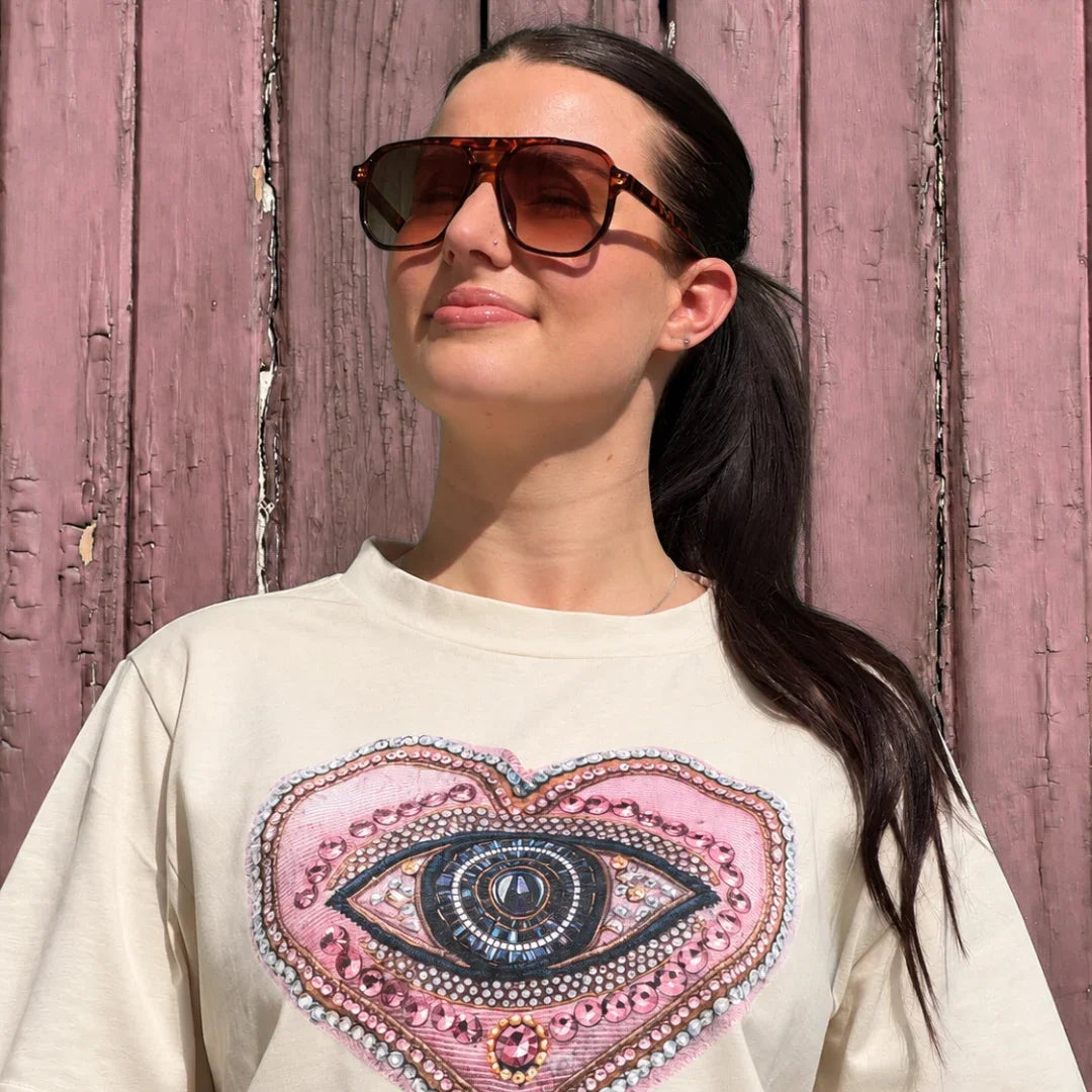 A woman wearing a cream-colored t-shirt with a large pink heart-shaped graphic featuring a detailed eye design, posed against a rustic pink wooden wall.