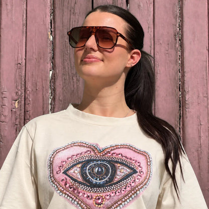 A woman wearing a cream-colored t-shirt with a large pink heart-shaped graphic featuring a detailed eye design, posed against a rustic pink wooden wall.