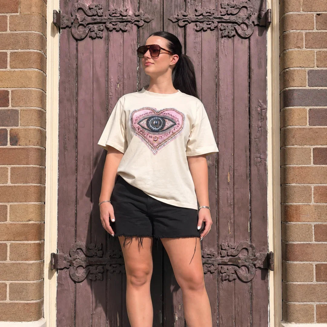 A woman stands in front of a rustic wooden door wearing a cream graphic t-shirt with a pink and blue eye design, black shorts, and white sneakers.