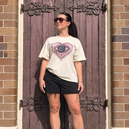 A woman stands in front of a rustic wooden door wearing a cream graphic t-shirt with a pink and blue eye design, black shorts, and white sneakers.
