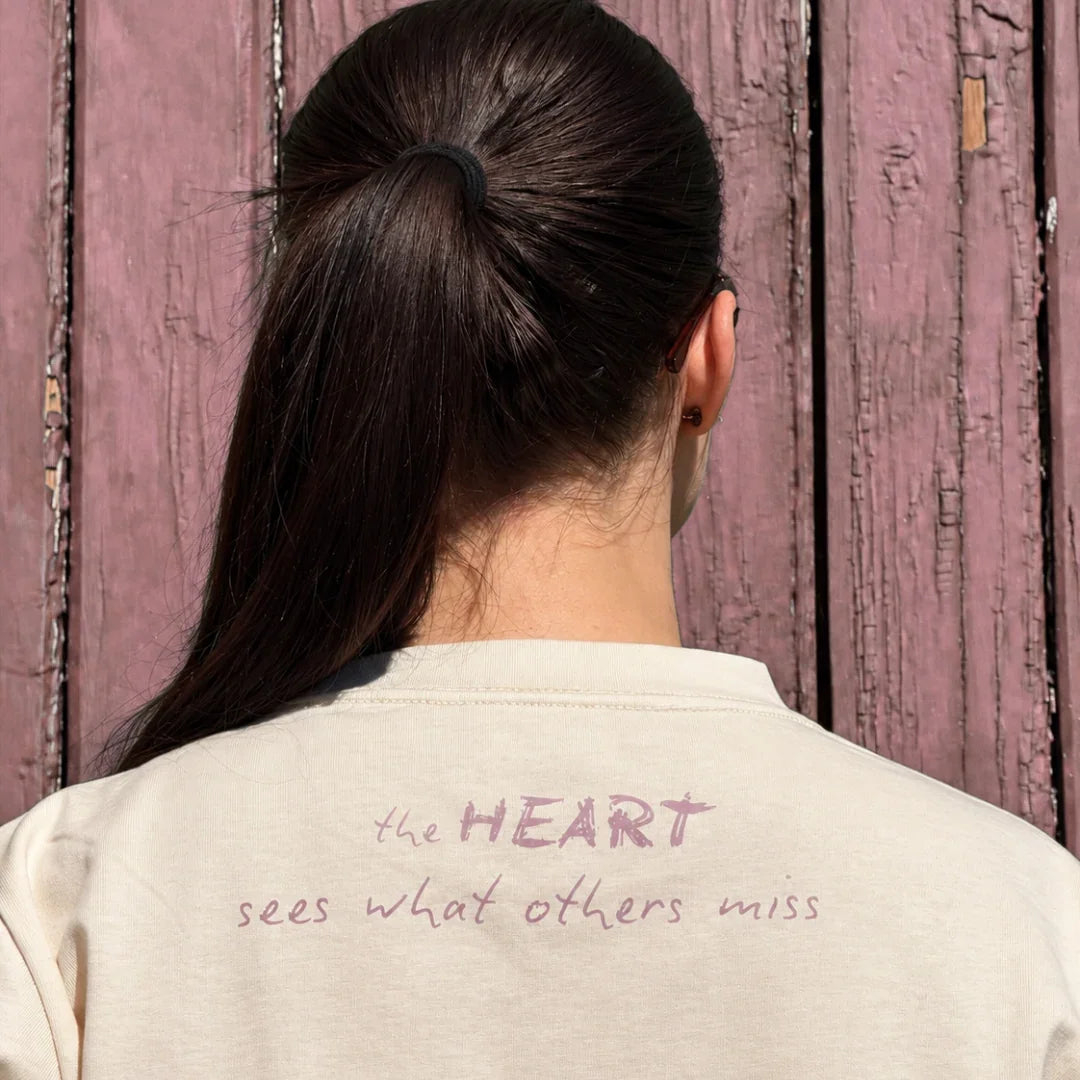 A person viewed from behind wearing a cream-colored crewneck sweatshirt with the phrase 'the HEART sees what others miss' printed in dusty pink on the back, set against a rustic pink wooden door.