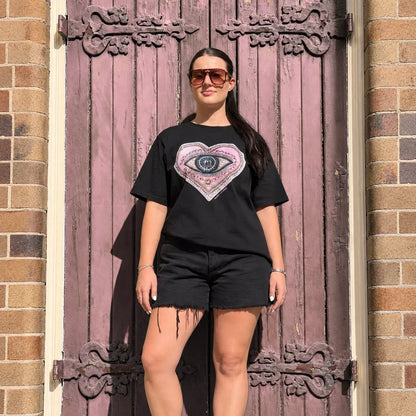 Woman wearing Bespoke Hearts The Lover’s Eye black tee and denim shorts by a rustic purple door.